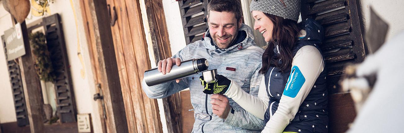Pärchen sitzt vor einer Hütte und schenkt sich Tee aus der Thermosflasche ein